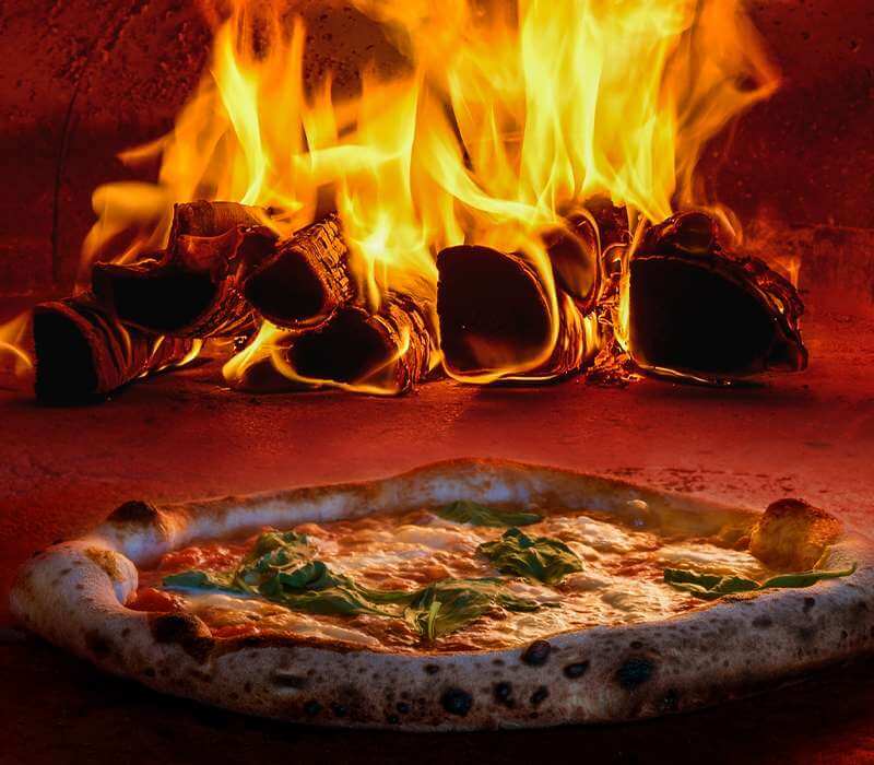 A Neapolitan-style pizza with basil in front of burning logs in a wood-fired oven