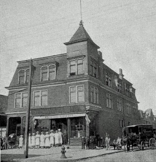 Historic photograph of an early bakery building on a street corner, with staff in white aprons lined up at the storefront and a horse-drawn wagon at the curb
