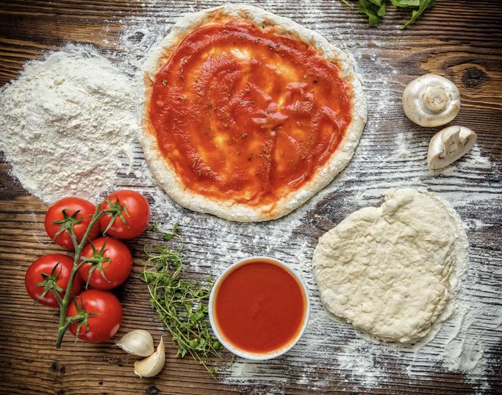 Sauced pizza dough, a stretched dough ball, flour, tomatoes, thyme and garlic on a floured wooden table